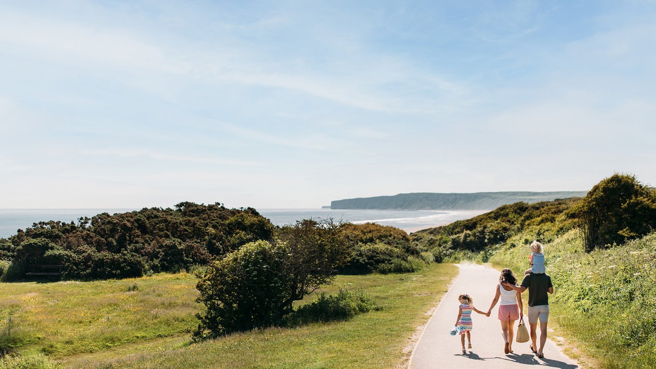 
              A young family are walking along a dry dirt path which is situated between rolling green hills on a hot summer's day.
              